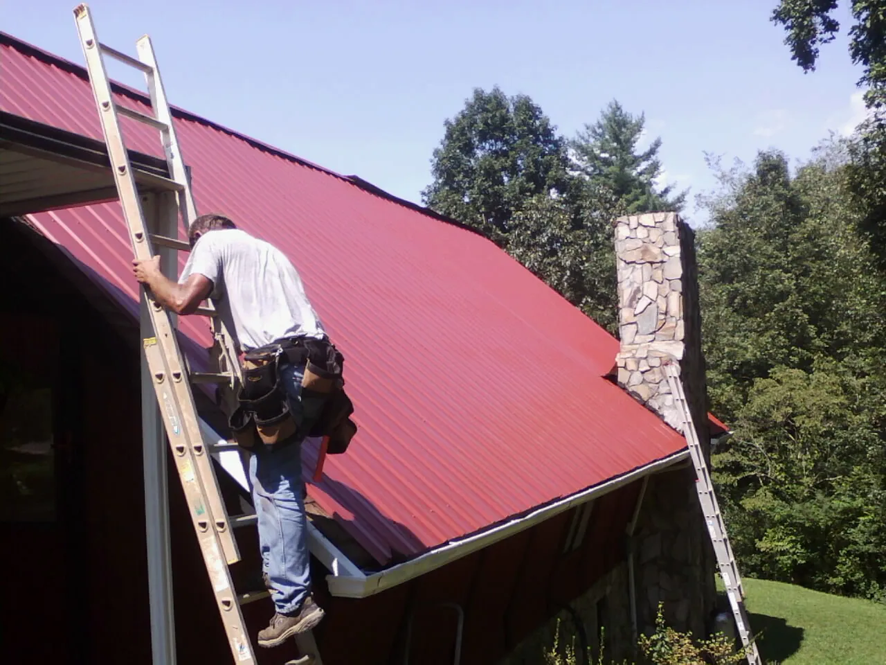 Roofer on a ladder beside a red metal roof and stone chimney — Soffit & Fascia Repair by Roofing Brothers in Saugerties